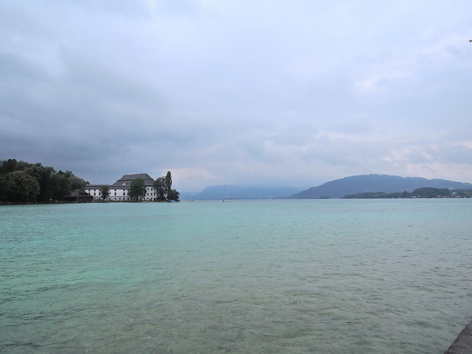 Der Attersee mit türkisfarbenem Wasser, am Ufer ein Gebäude, im Hintergrund die Berge des Salzkammerguts.