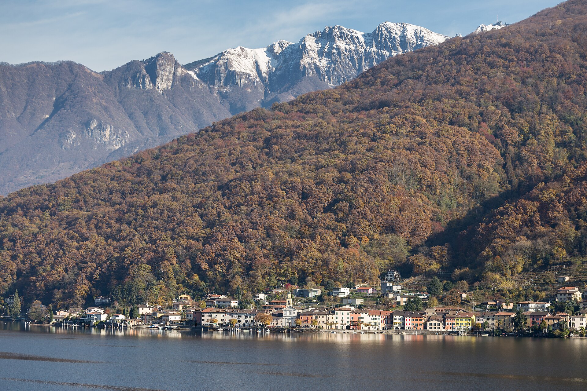 Ein Dorf am Ufer des Luganersees, dahinter bewaldete Hänge und verschneite Gipfel
