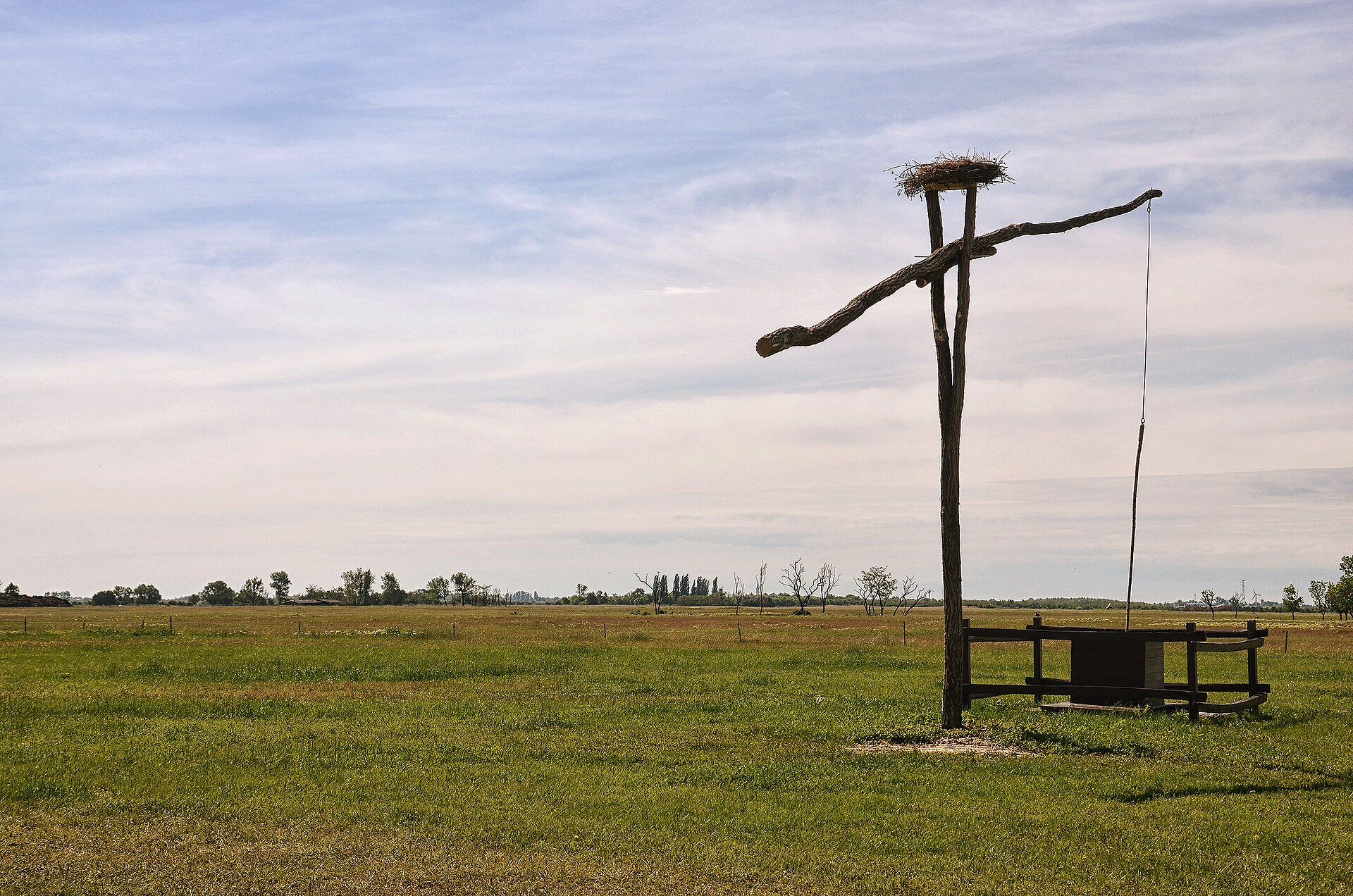 Weite, gehölzarme Puszta-Ebene in Ungarn; rechts ein hölzerner Ziehbrunnen mit Storchennest auf dem Galgen