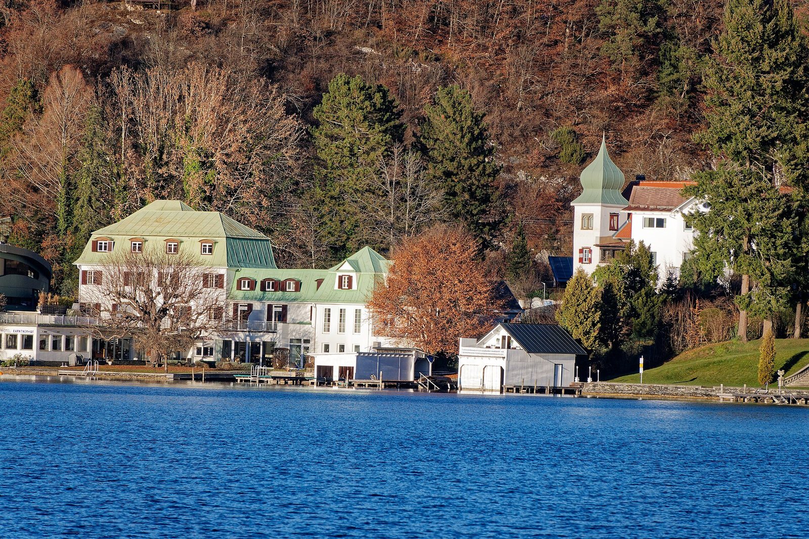 Das Ufer des Wörthersees bei Pörtschach mit Häusern direkt am Wasser und einem herbstlich gefärbten Hang dahinter.